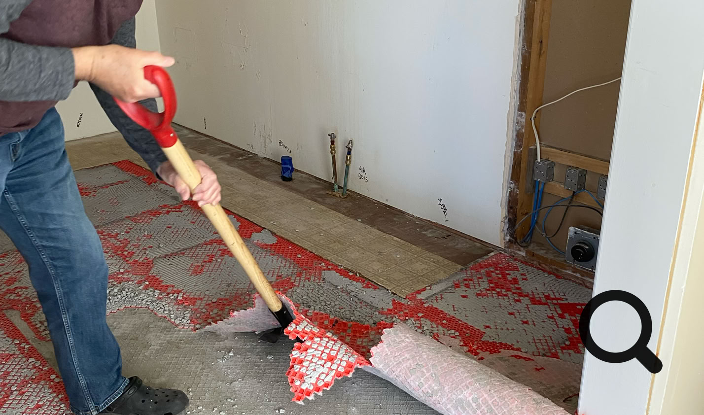 A person using a shovel to remove red material from a floor in a room under renovation, with exposed wall sections.