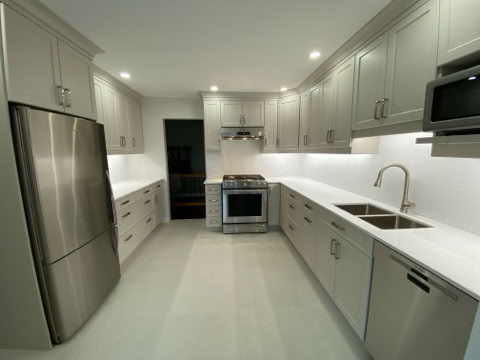 Modern kitchen with gray cabinets, stainless steel refrigerator, oven, microwave, and a double sink. White countertops and recessed lighting are present.