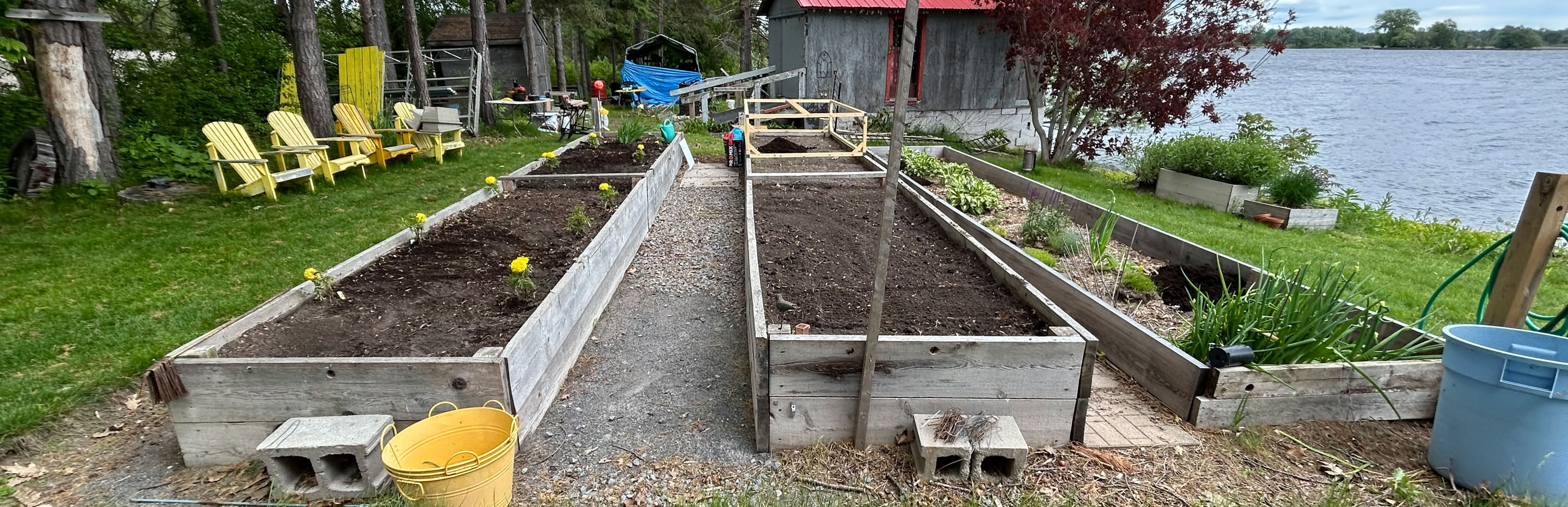 Raised garden beds with soil, surrounded by grass and trees, with a red-roofed building and a body of water in the background.