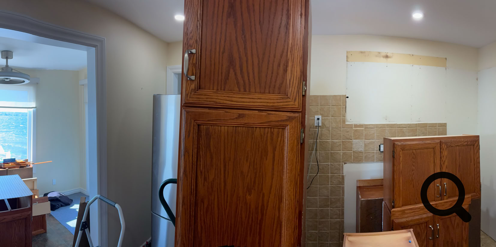 Panoramic view of a kitchen under renovation, featuring wooden cabinets, a window with a curtain, a ceiling light, and construction materials scattered around.