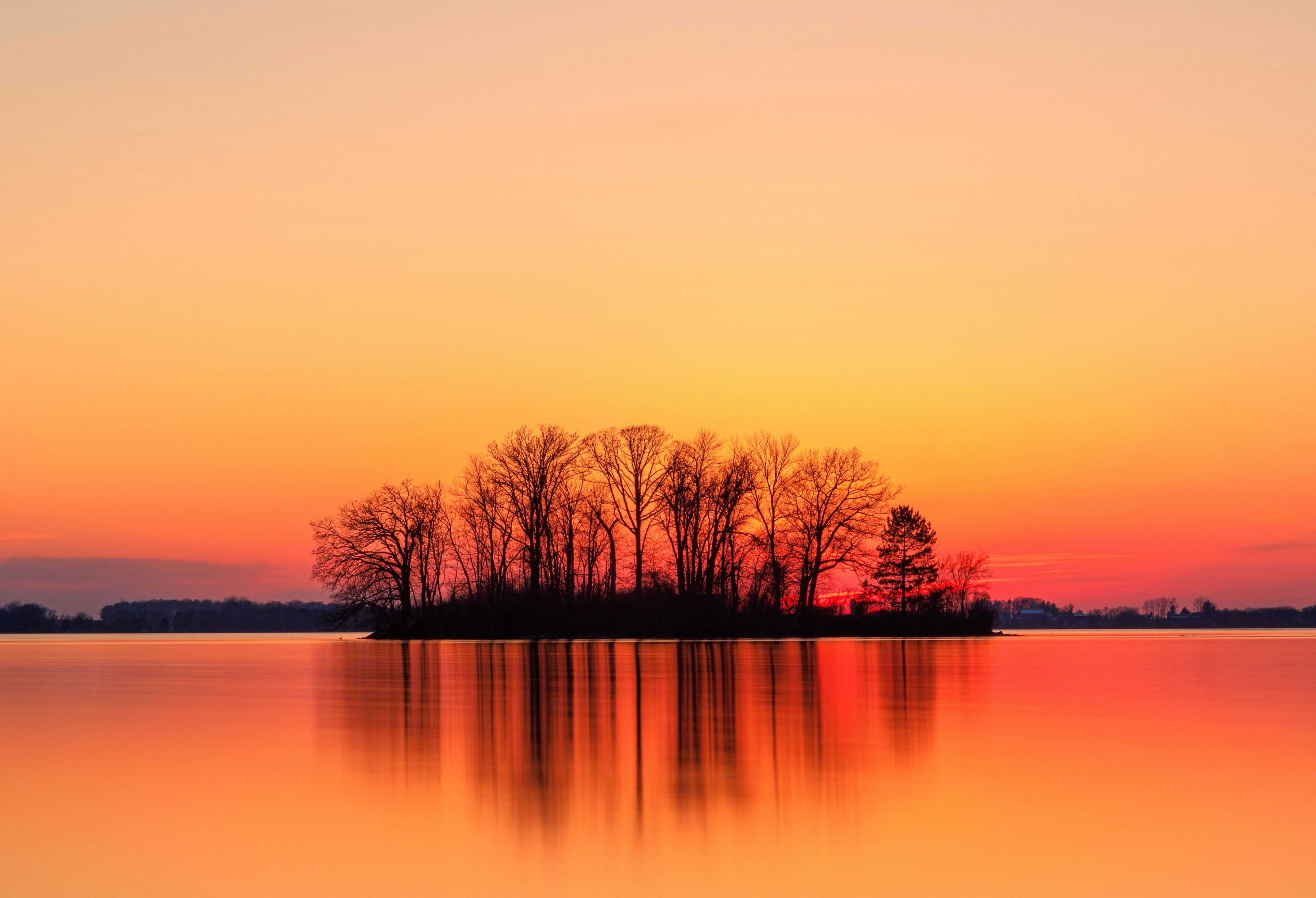 A small island with bare trees is reflected in calm water during a vibrant sunset, with an orange and pink sky.